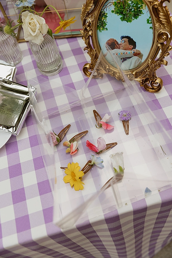 Wedding flat lay bridal detail flat lay with ornate gold frame photo, floral hair clips, and colorful blooms on a purple gingham tablecloth