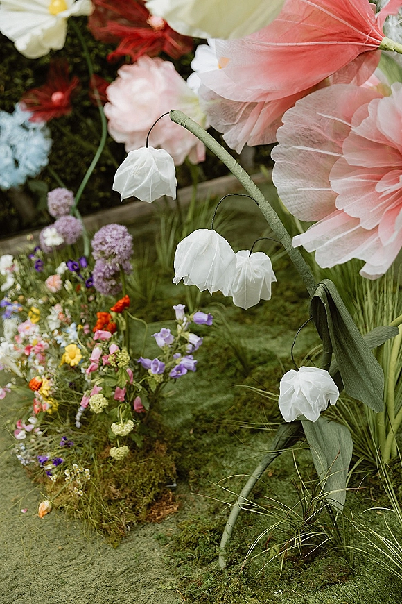 Wedding floral installation with oversized paper flowers and hanging flower lanterns over a mossy wildflower meadow on an indoor floor