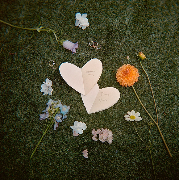 Wedding vows in heart vow booklet with handwritten pages, gold wedding bands, and scattered flower stems on a grass lawn flat lay