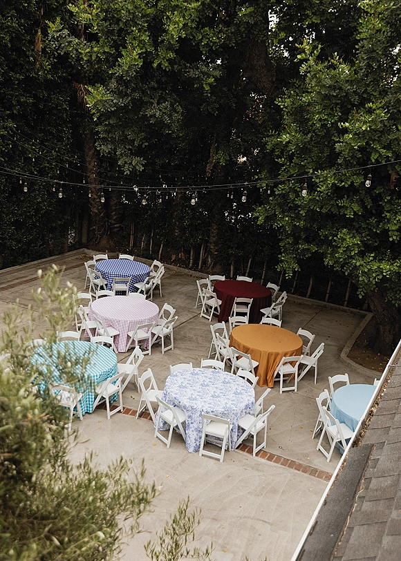 Outdoor reception setup with backyard wedding reception round tables in colorful tablecloths, white folding chairs, and string lights under a tree canopy courtyard patio