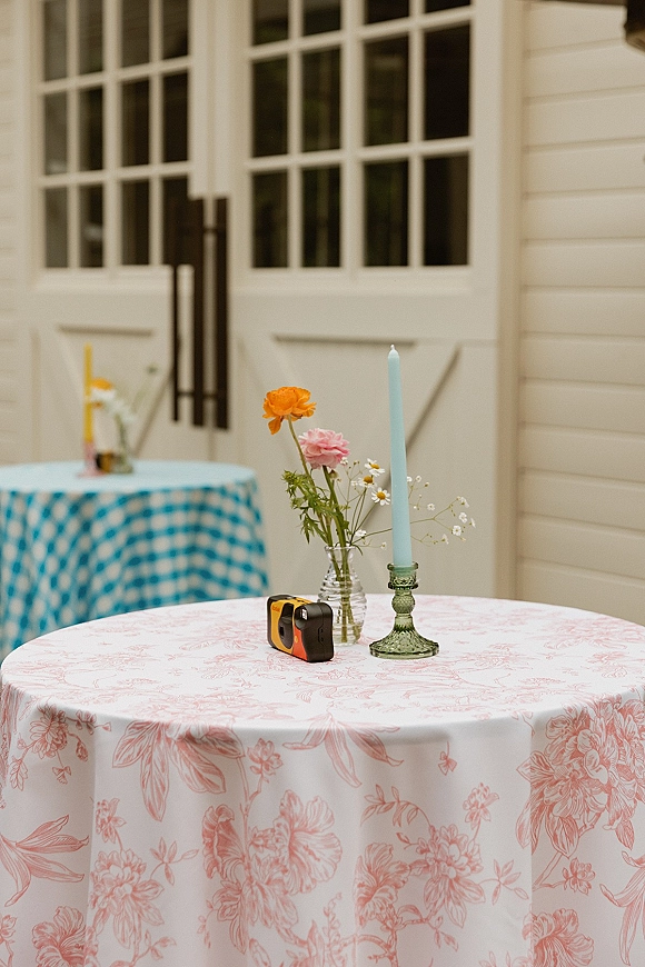 Reception table decor with a wedding bud vase centerpiece of wildflowers beside a taper candle on a gingham tablecloth by white barn doors