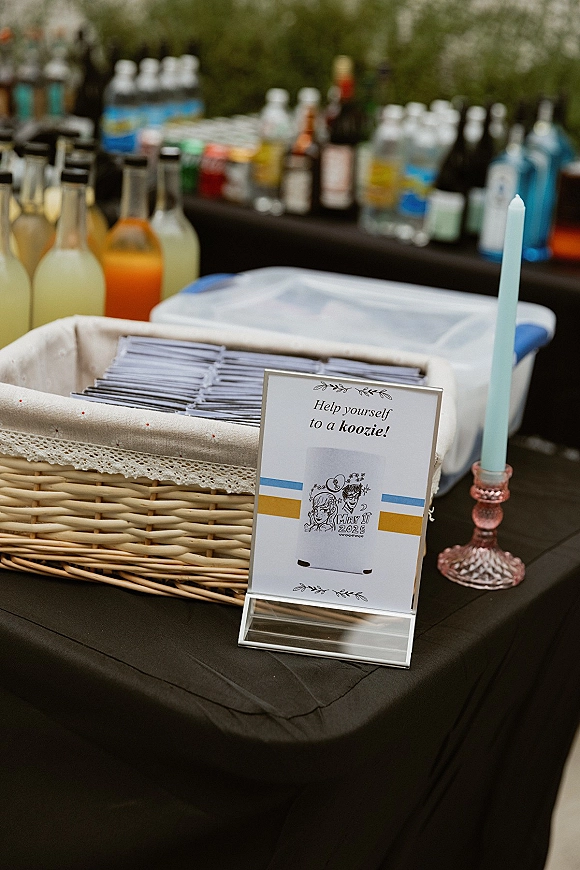 Wedding koozie display with custom wedding koozies in a lace-trim wicker basket, acrylic help-yourself sign and bottled drinks on bar table outdoors
