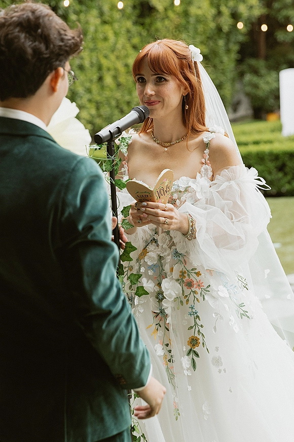 Wedding vows as the bride reading vows into a microphone, veil and floral embroidered dress visible, with garden hedges and string lights behind
