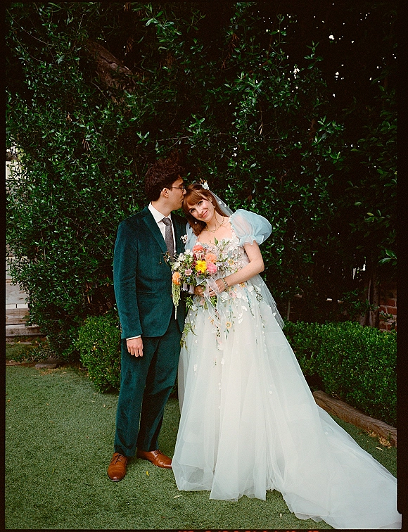 Couple portrait of groom kissing bride’s forehead as she holds a colorful bouquet, in a puff sleeve gown before garden hedges and brick wall