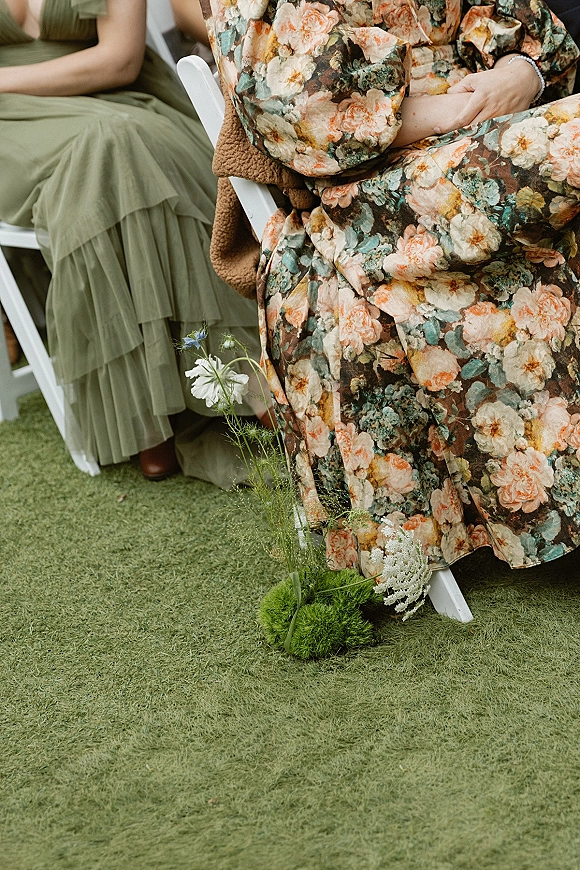 Wedding guest attire featuring a floral wedding guest dress with a bracelet beside white folding chairs and mossy florals on a grass lawn