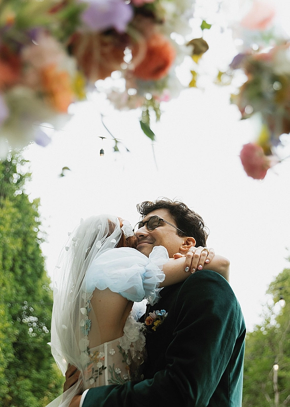 Couple portrait of bride and groom in a newlywed hug under a hanging floral canopy, bride’s polka-dot veil and appliqué dress visible