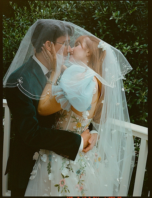 Wedding kiss portrait of bride and groom kissing under a veil, bride touching his face with rings visible against green foliage and white railing