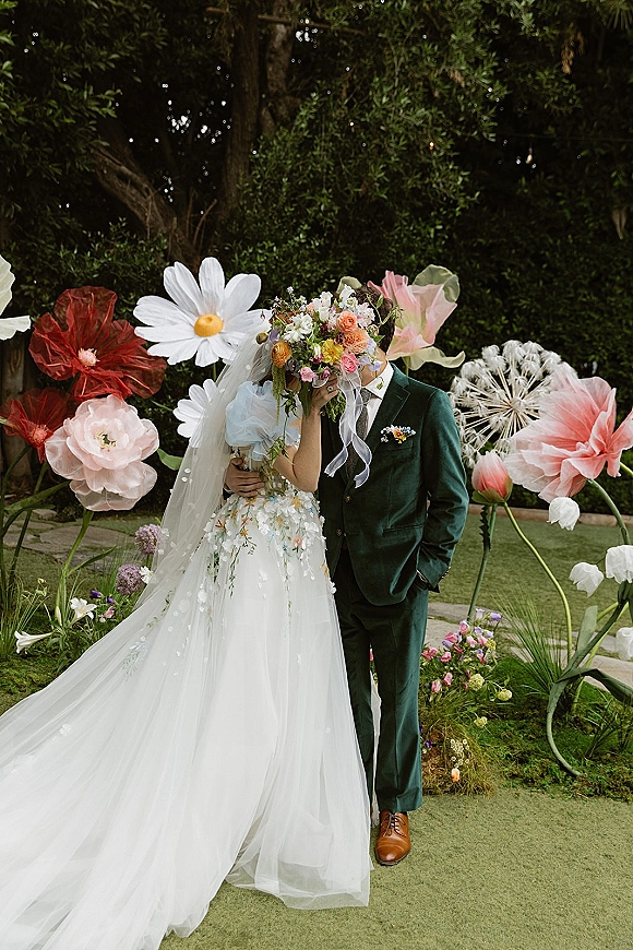 Couple portrait of bride and groom pose, bride hiding behind a colorful bouquet with veil, beside oversized paper flowers in a garden