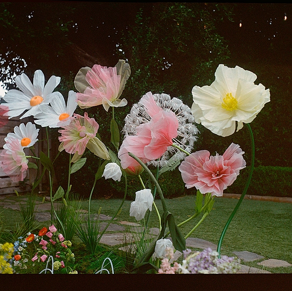 Floral wedding decor featuring giant paper flowers decor with oversized blooms and stems along a garden lawn by a stone path, under string lights