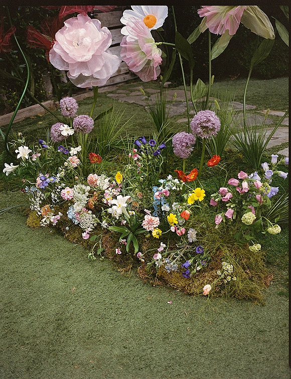 Wedding floral installation with wildflower ceremony decor on a moss base, oversized paper flowers and purple alliums by a stone path on grass lawn