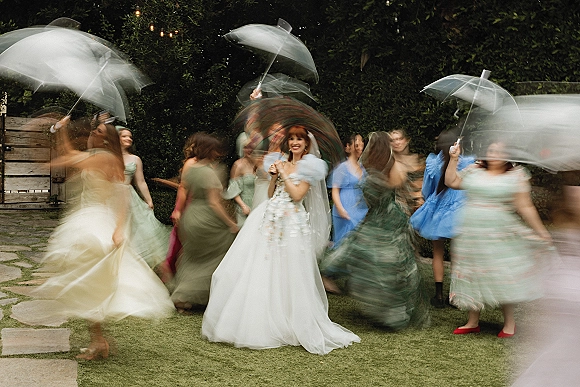 Bride portrait with bridesmaids holding parasols under string lights, walking along a stone path by a garden hedge and wooden gate