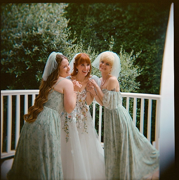 Bridesmaid portrait of three women in green floral dresses and veils, one in an embroidered gown, smiling on a porch with foliage behind