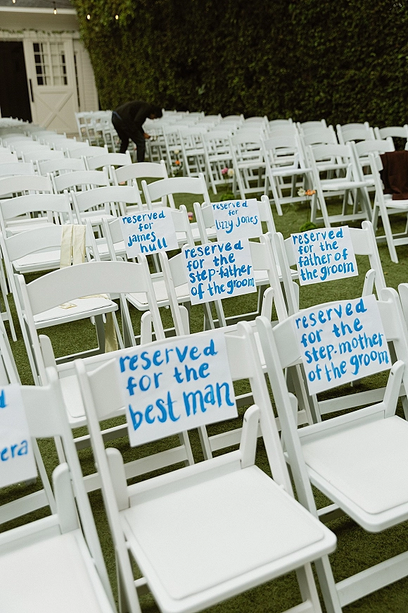 Ceremony seating signs with blue lettering tied by ribbon to white folding chairs, marking reserved seats on a grass lawn by a hedge wall