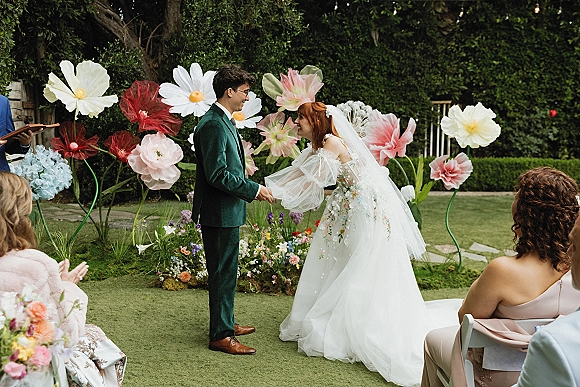 Wedding vows as bride and groom hold hands facing each other, bride in embroidered off-shoulder gown and veil before paper flowers.