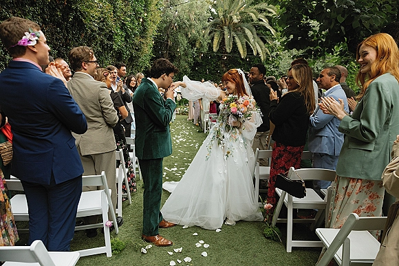 Ceremony recessional as newlyweds walking down aisle, bride with cathedral veil and colorful bouquet, guests applauding beneath string lights in garden