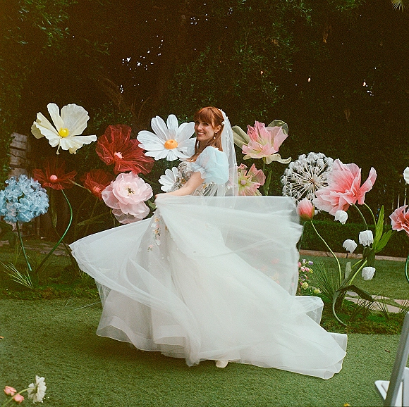 Bridal portrait of a bride twirling dress with veil in motion, tulle skirt flowing by a garden hedge and oversized paper flowers backdrop
