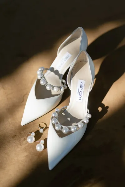 Bridal shoes with pearl ankle straps beside pearl earrings and a diamond engagement ring on tan fabric with sunlit shadows