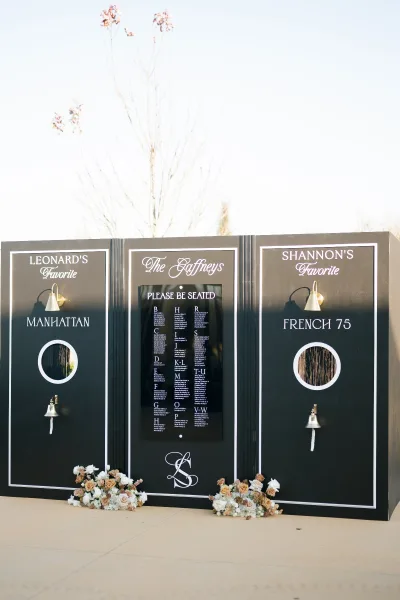 Wedding seating chart on a black calligraphy board with monogram initials, brass sconces and service bells, set outdoors with floral accents