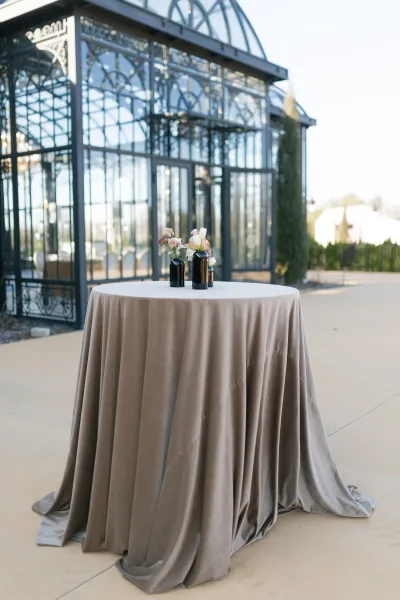 Cocktail table decor with taupe linen on a wedding cocktail table, styled with black bud vases and a small floral arrangement in a glass conservatory setting