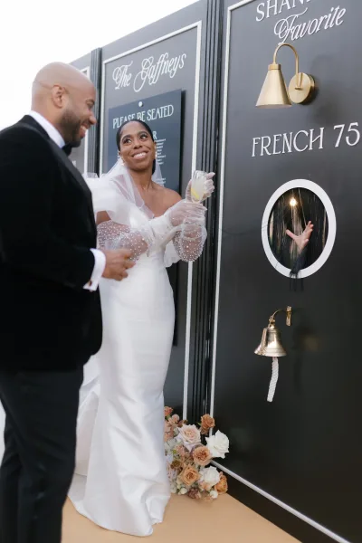 Wedding champagne toast as bride in gloves and veil clinks coupe glasses with groom in tuxedo before black-and-white signage wall and lemon twist garnish