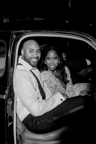 Couple portrait of newlyweds in car, bride in beaded lace wedding dress and pearl earrings beside groom in bow tie at night street