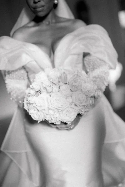 Bridal portrait in black and white of a bride holding bouquet of white roses, wearing a strapless pearl-beaded dress with veil and earrings indoors