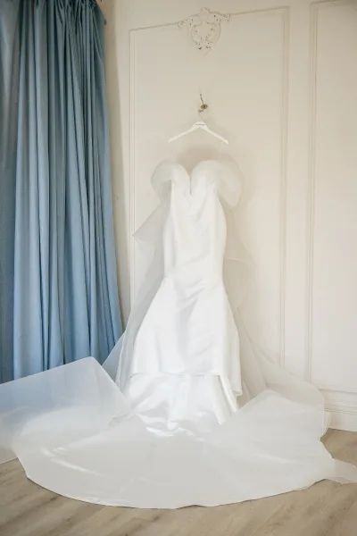 Wedding dress hanging on a hook, a strapless wedding dress with a tulle overskirt against a white paneled wall and blue curtains