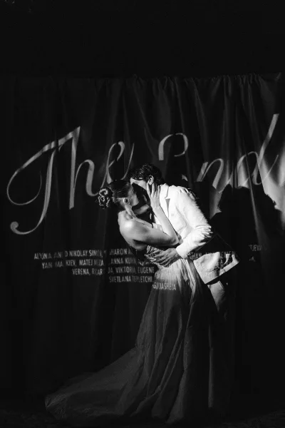 Wedding kiss portrait in black and white with dramatic spotlight shadows as groom holds bride in strapless tulle gown before lettered curtain backdrop