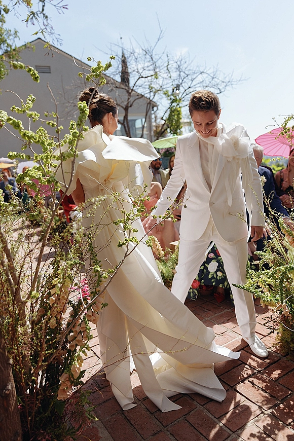 Recessional moment as bride and groom walking aisle, her oversized bow gown and long train trailing past cheering guests with umbrellas in a brick courtyard