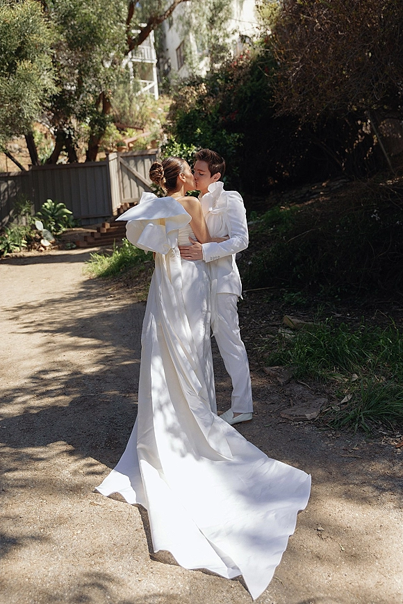 Wedding kiss portrait of bride and groom kissing on a tree-lined outdoor path, her long train and oversized bow trailing behind