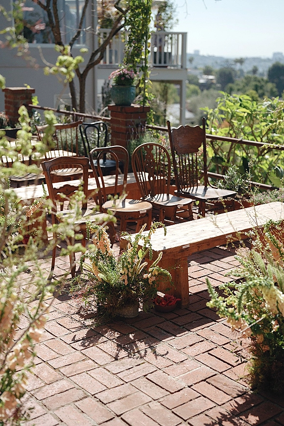 Ceremony seating with mismatched chair wedding ceremony lineup, benches and aisle florals on brick pavers, framed by patio trees and railing