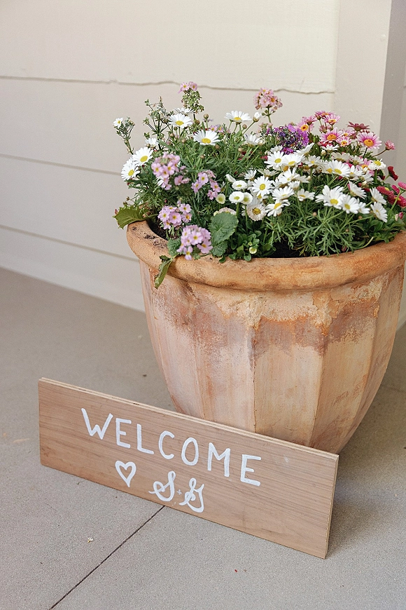 Wedding welcome sign with hand painted lettering and a heart doodle, leaning by a terracotta planter of daisies against a white wall