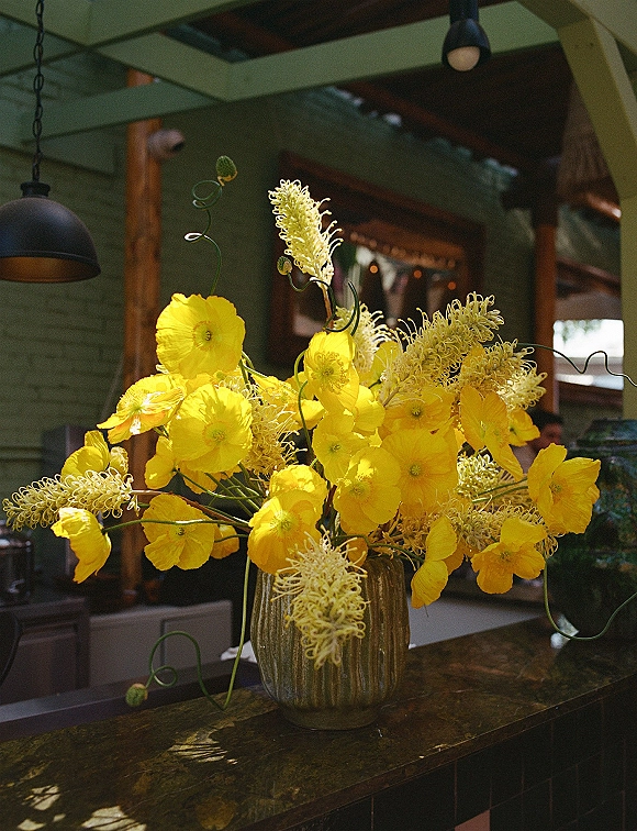 Floral centerpiece of bright yellow flowers in a ceramic vase with curly greenery stems on a bar counter beneath pendant lights