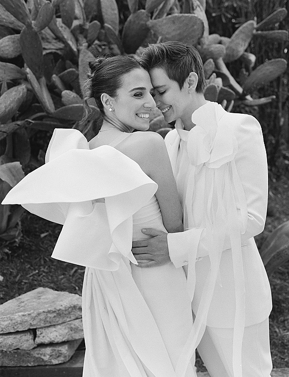 Couple portrait of two brides embracing with a forehead touch, one in a ruffled one-shoulder gown, in a cactus garden by a stone bench