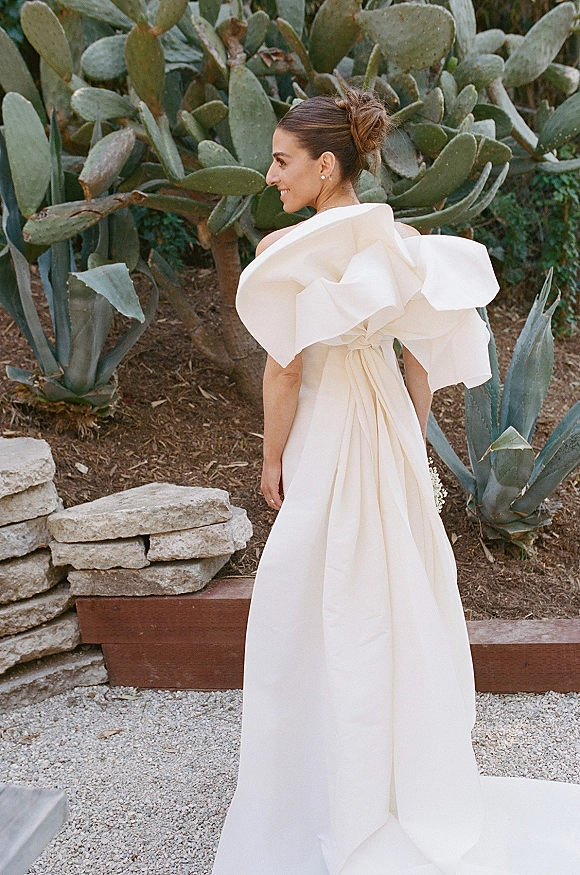 Bridal portrait of a bride in a back view wedding dress with an oversized bow and draped train, walking a cactus garden gravel path