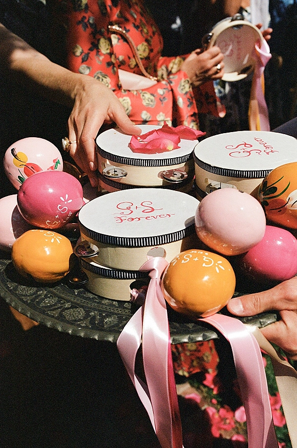 Wedding tambourines with satin ribbons and calligraphy lettering held up by guests in a dark indoor crowd, with rose petals and maracas