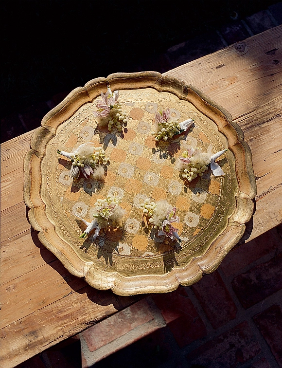 Wedding boutonnieres arranged as a boutonniere set with dried florals and white ribbon wrap on an antique gold tray in sunlit shadows