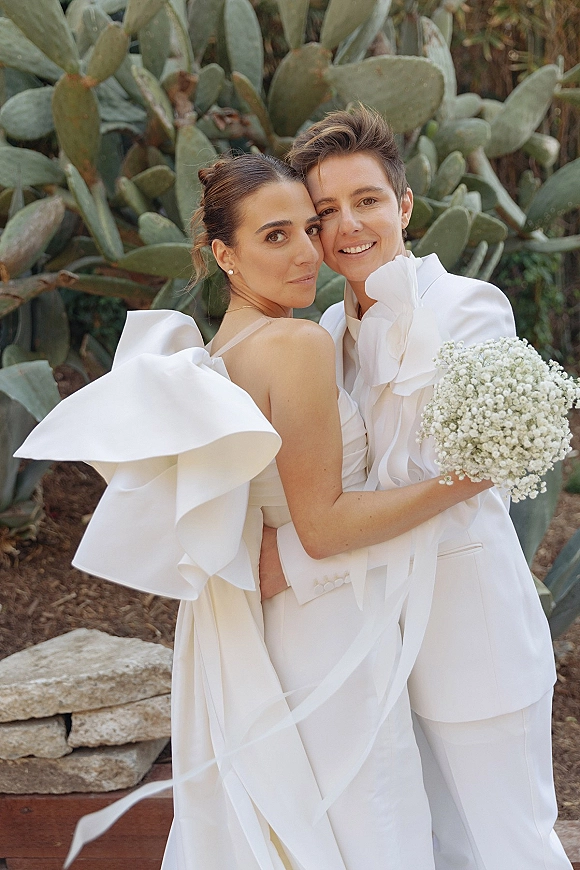 Couple portrait of a same sex wedding couple hugging, one in a ruffle sleeve dress and one in a white suit holding a baby's breath bouquet in a cactus garden