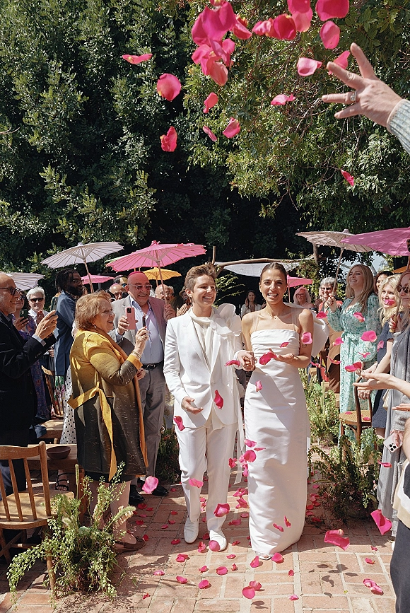 Wedding recessional as newlyweds walk the brick garden aisle through a rose petal toss, guests cheering under parasols and greenery