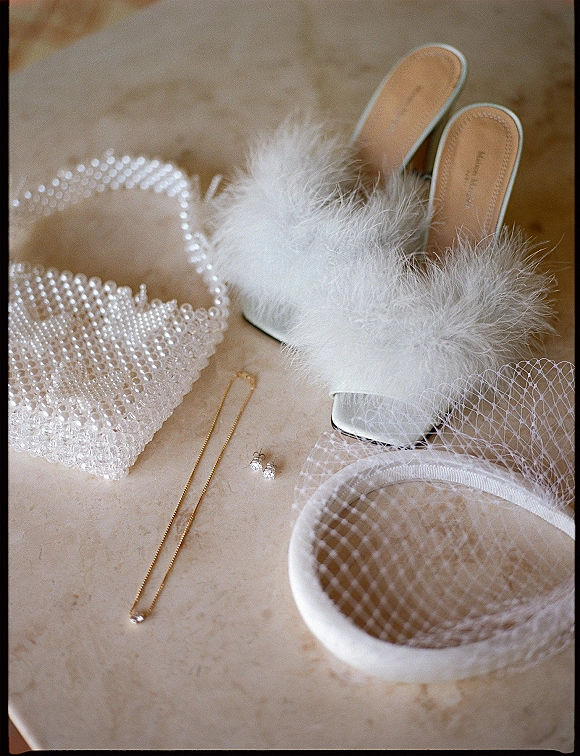 Bridal accessories flatlay featuring white feather mules, pearl beaded handbag, birdcage veil headband and gold pendant on stone tabletop