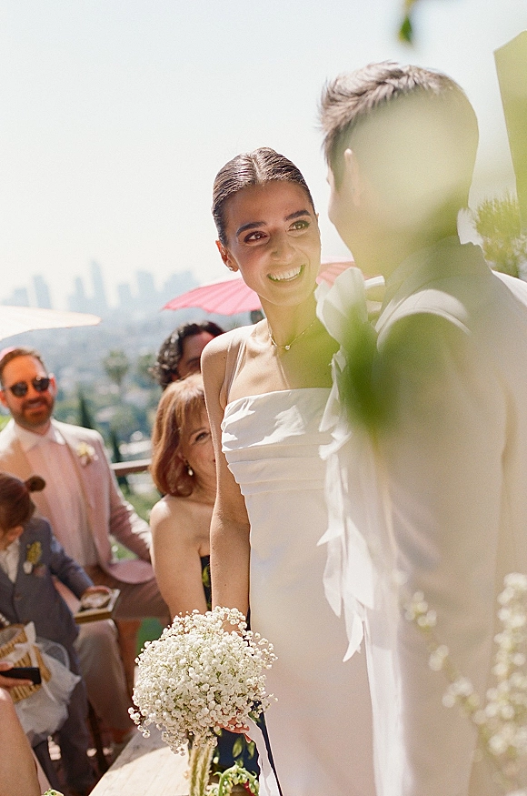 Ceremony moment as bride smiles at groom, holding a baby's breath bouquet in a strapless dress on a terrace with skyline and parasol guests