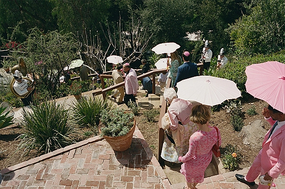 Wedding processional with outdoor wedding processional guests carrying parasols as a brass band leads down a garden staircase to a brick walkway