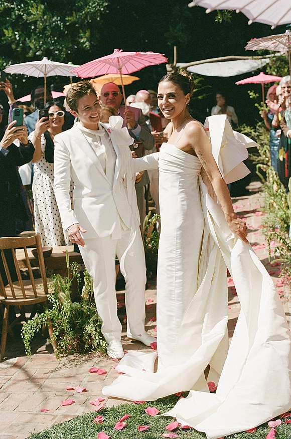 Recessional moment as newlyweds walking down aisle, one in a white gown with bow train and one in white suit, guests toss rose petals on brick path