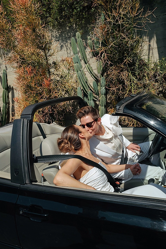 Wedding couple portrait of bride and groom in car wearing sunglasses, holding hands in a sunlit convertible beside cacti and stucco wall