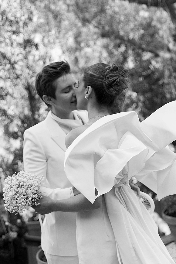 Wedding kiss portrait of bride and groom kissing, groom in white suit and bride in ruffle-sleeve gown holding bouquet in garden setting