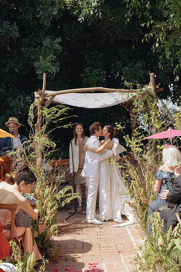 Ceremony kiss beneath a fabric-draped wedding arch canopy with greenery, rose petals on a brick aisle, and guests in a garden courtyard