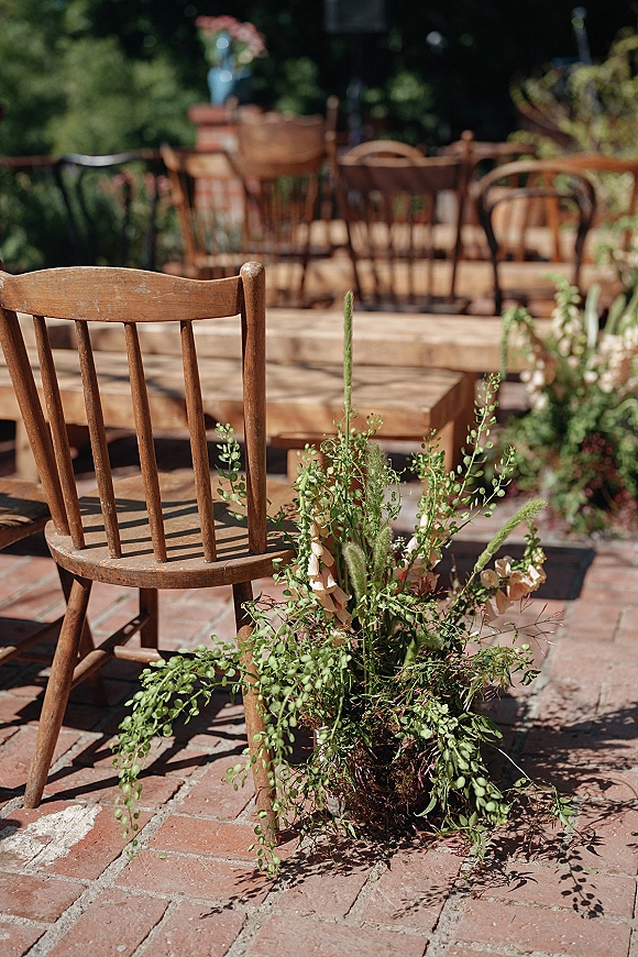 Reception seating with wooden wedding chairs around long farm tables, greenery and a ground floral arrangement on a sunlit brick patio