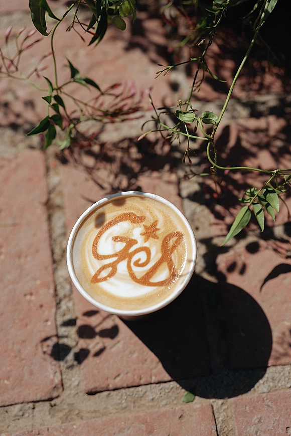 Wedding latte art in a paper cup with a wedding monogram latte foam design on brick pavement with leafy shadow patterns