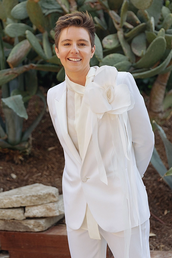 Groom portrait in a white wedding suit with a neck scarf, posing by a cactus garden with an oversized fabric flower corsage on shoulder