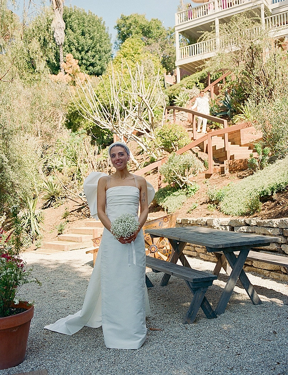 Bridal portrait of a bride in a strapless wedding dress with bow detail, birdcage veil, and baby’s breath bouquet in a garden setting